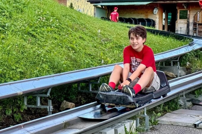 A person enjoying a luge ride on a metal track winding through a green hillside, with a luge station visible in the background.