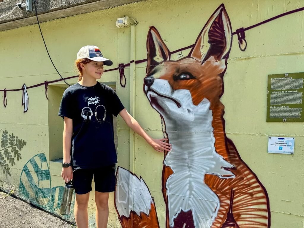 A person in a baseball cap poses right next to a large, vibrant mural of a fox painted on a pale yellow wall.