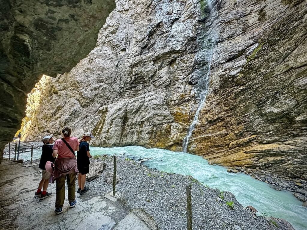 Trailside view of a vibrant turquoise stream flowing through a rocky canyon with a cascading waterfall.