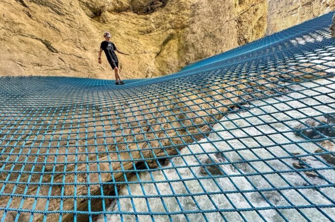 A person walks on a blue metal grid bridge suspended against a rocky cliff face.