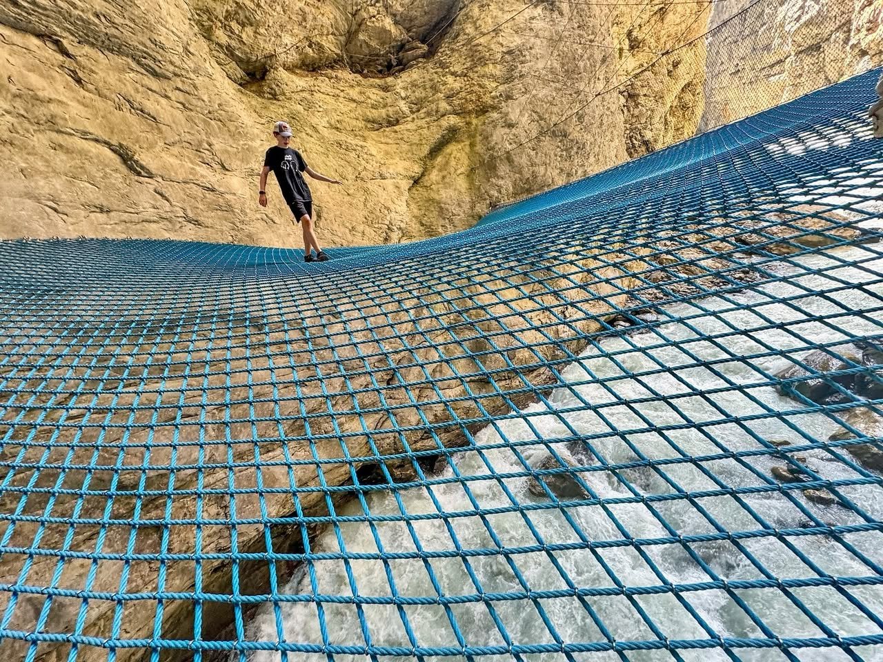 A person walks on a blue metal grid bridge suspended against a rocky cliff face.