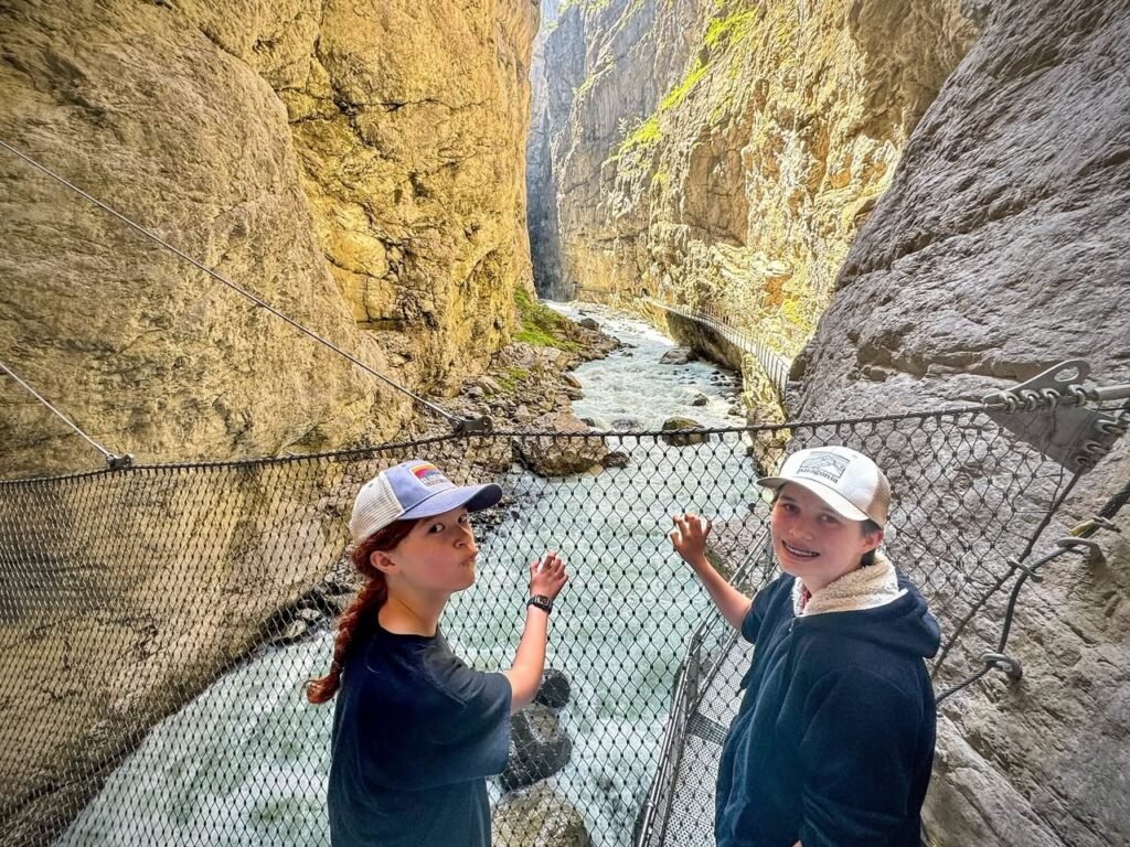 Two visitors standing on a metal mesh bridge inside the narrow Fjaðrárgljúfur Canyon, with a small stream flowing between towering canyon walls.