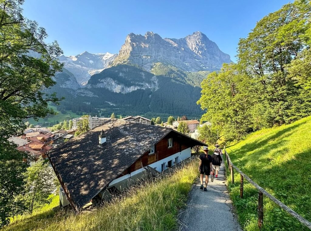A narrow path leads past a traditional red and white alpine house toward distant dramatic mountain peaks under a clear blue sky.