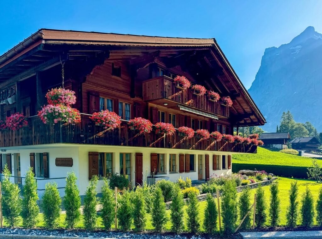 A rustic wooden chalet with red flower baskets adorning the balconies, set against a backdrop of lush greenery and towering mountains under a clear blue sky.