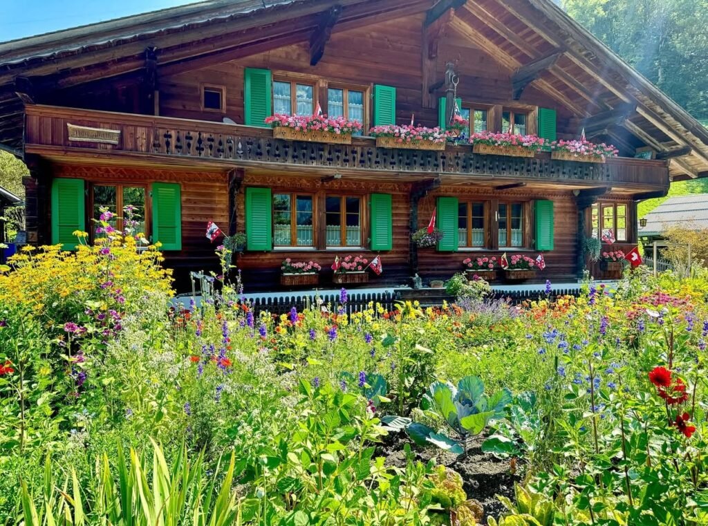 A rustic log cabin with green shutters and a balcony surrounded by a lush, colorful garden.