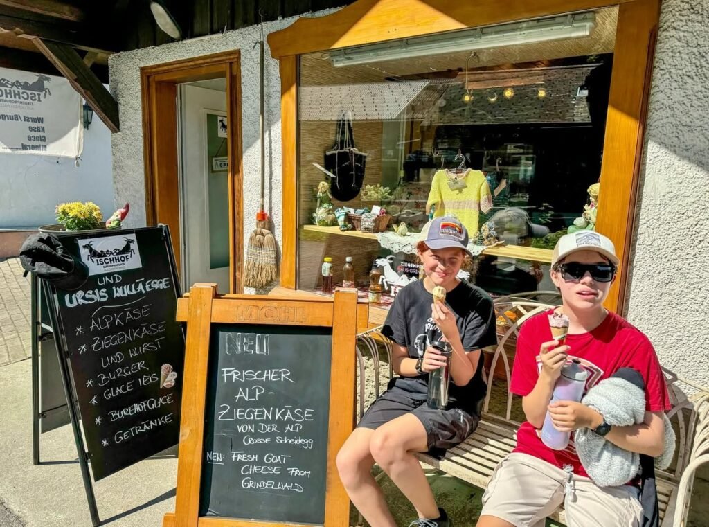 A family sitting on a wooden bench outside a cafe, eating ice cream and holding drinks. The scene features chalkboard menus with German text and a bright, sunny atmosphere.