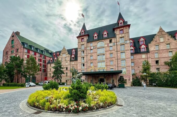 A grand chateau-style hotel features a cobblestone entrance with a vibrant circular garden full of yellow flowers and evergreen shrubs.