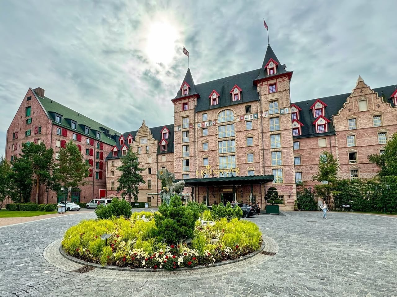 Hotel Krønasår A grand chateau-style hotel features a cobblestone entrance with a vibrant circular garden full of yellow flowers and evergreen shrubs.