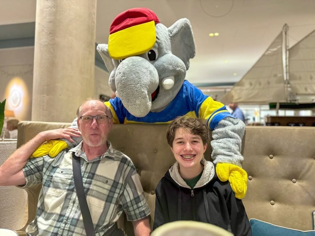 Two people smiling and posing with a colorful elephant mascot in a lounge setting. The scene highlights a friendly atmosphere and travel enjoyment.