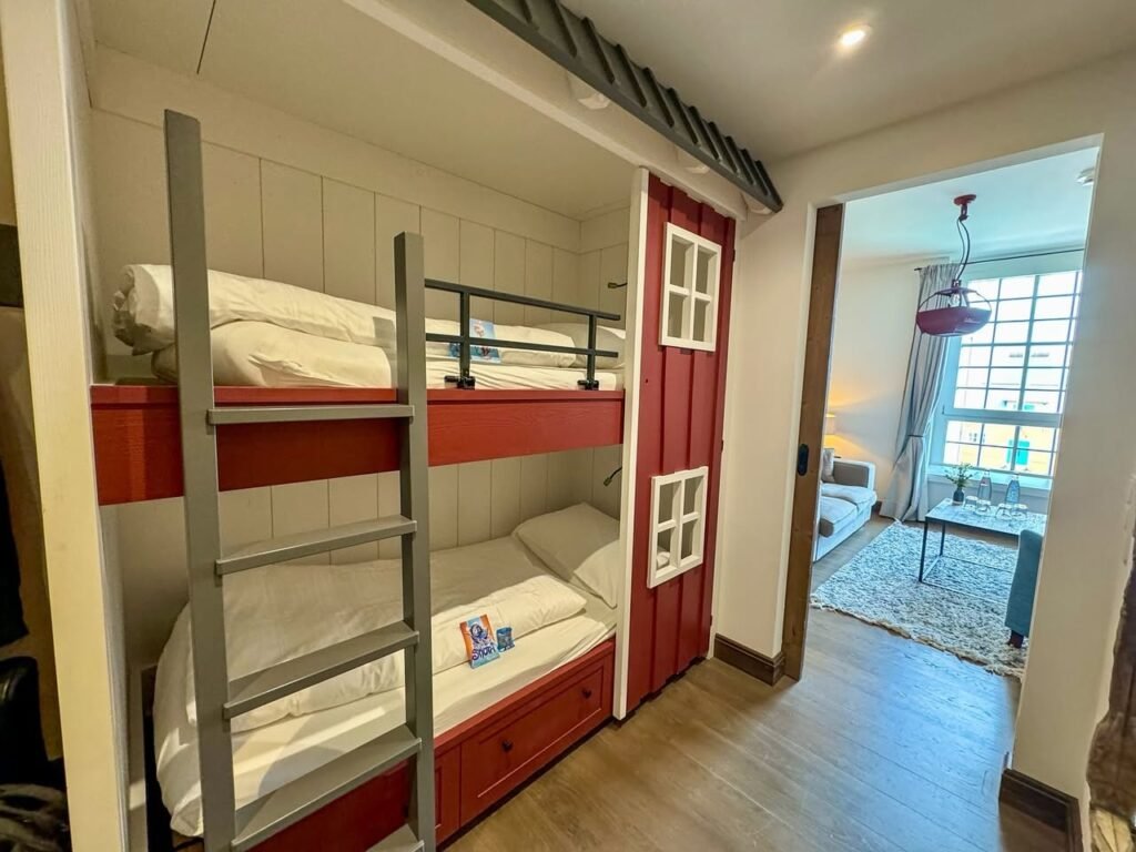 A bright bedroom featuring red bunk beds with stairs and storage beneath. Natural light streams in from a nearby dining area.