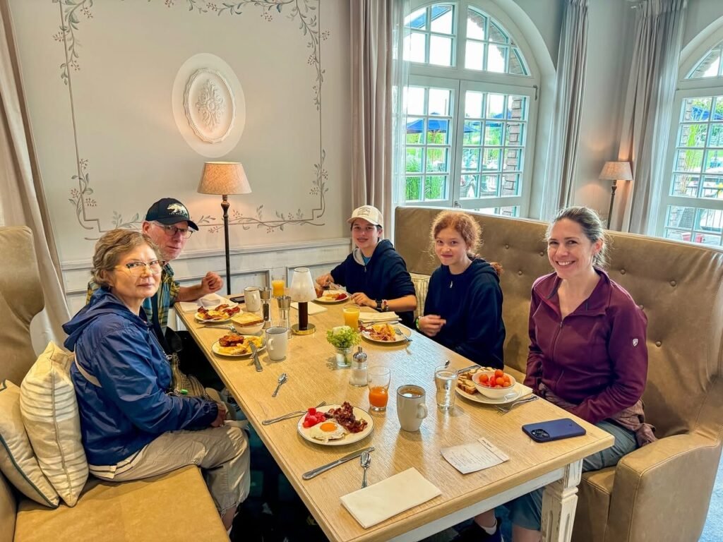 A family gathered for a shared meal in a bright dining room with arched windows and comfortable seating. Plates of food and drinks are set on a wooden table in a cozy living space.