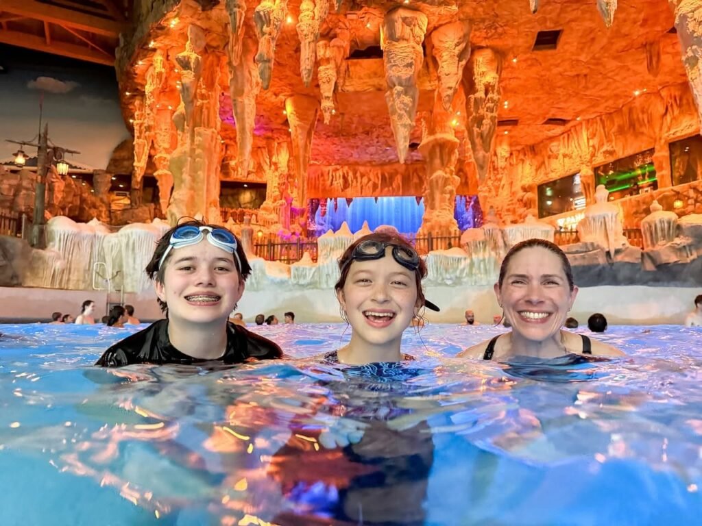 Visitors enjoying warm waters in a hot spring surrounded by an illuminated ice-like cave interior, featuring textured walls and colorful lighting.