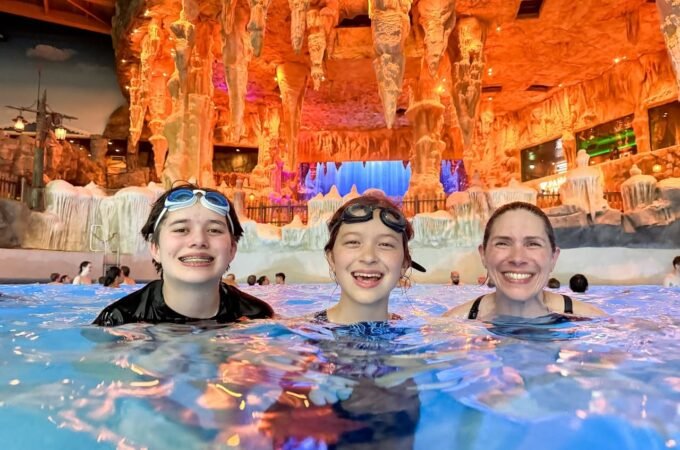 Visitors enjoying warm waters in a hot spring surrounded by an illuminated ice-like cave interior, featuring textured walls and colorful lighting.