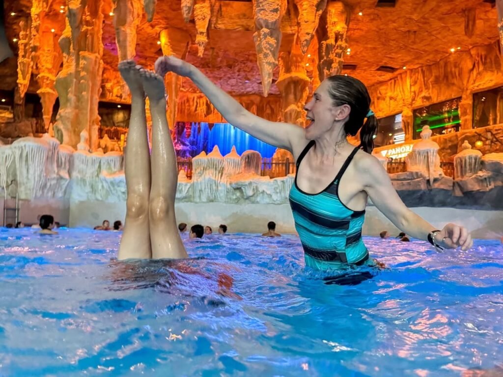 Person floating upside down in a bright blue indoor pool. Background features illuminated rock formations and a tropical atmosphere.
