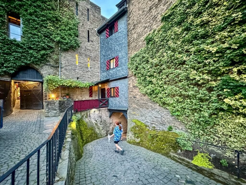 Cobbled path leading through an ivy-covered stone estate with red-shuttered turrets.