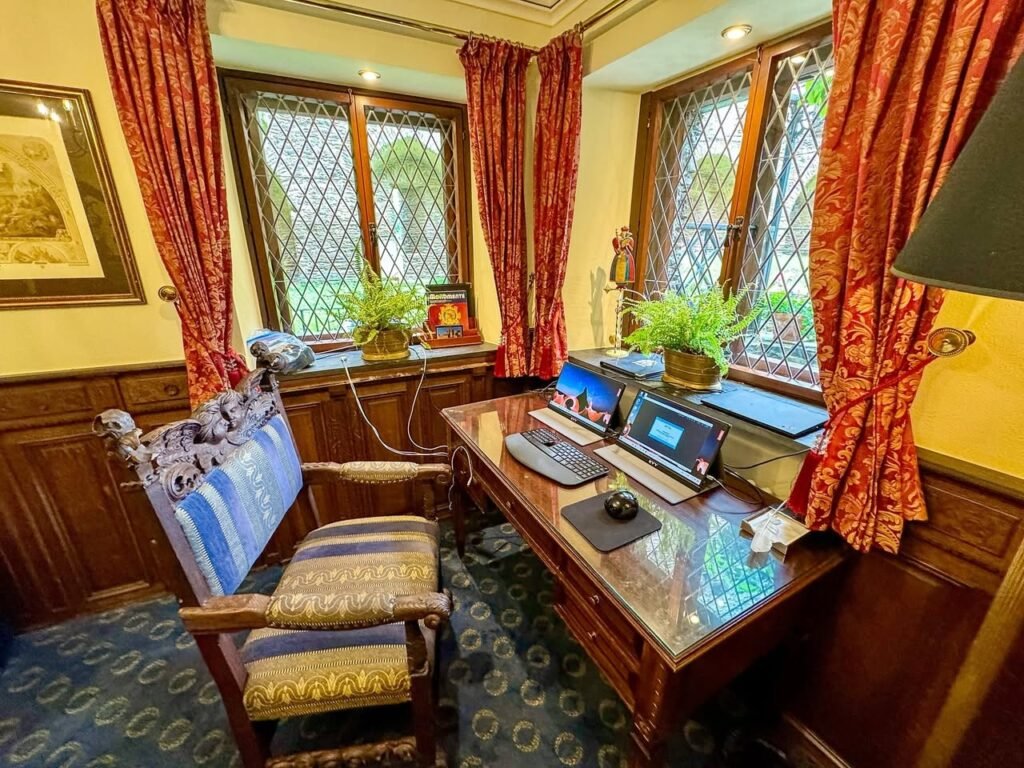 A beautifully appointed writing corner featuring a wooden desk with laptops nestled between red patterned drapes. The setting includes classic wood paneling and diamond-paned windows that add to its grand charm.