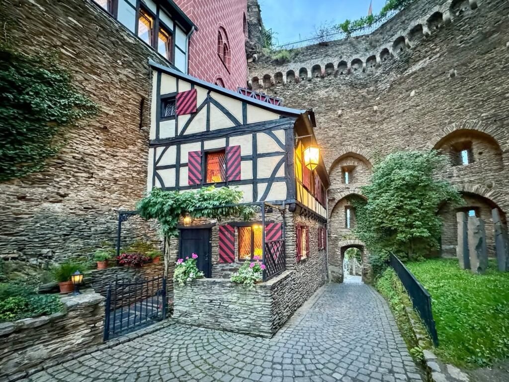 A cobblestone path winds past a traditional half-timbered cottage nestled directly into the massive rough-hewn stone wall of a castle.