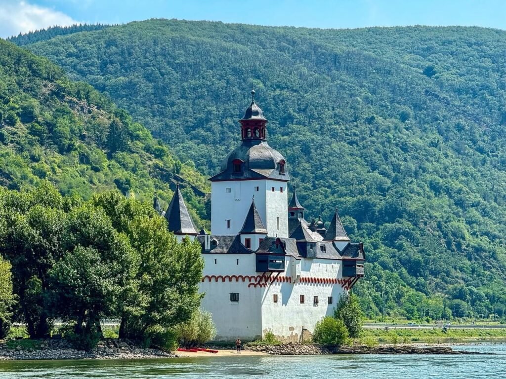 A white castle with dark turrets situated on a riverbank, surrounded by lush green mountains.