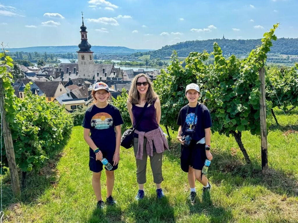 Three people walking through lush vineyards with medieval-style buildings and a prominent church steeple in the background under a blue sky.