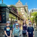 A family walks a cobblestone lane surrounded by charming half-timbered houses with hanging flowers. They stand near a decorative sign for a sausage shop.