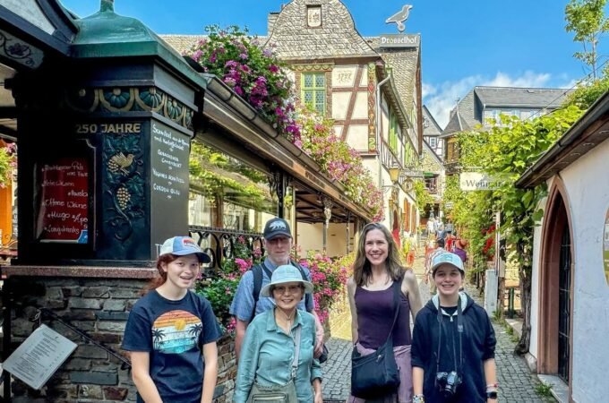 A family walks a cobblestone lane surrounded by charming half-timbered houses with hanging flowers. They stand near a decorative sign for a sausage shop.