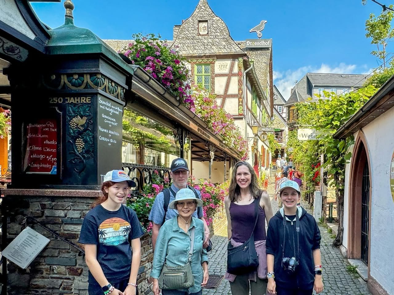 Drosselgasse Rüdesheim A family walks a cobblestone lane surrounded by charming half-timbered houses with hanging flowers. They stand near a decorative sign for a sausage shop.