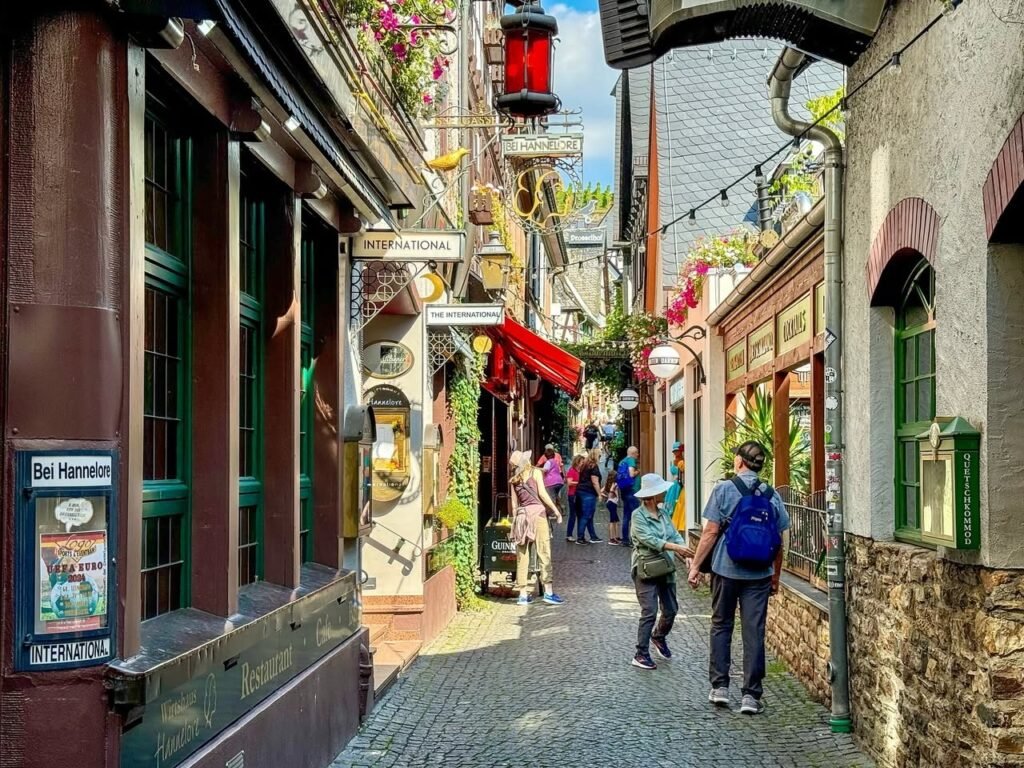 Visitors walk through a narrow cobblestone alley lined with historic buildings. Traditional architecture features charming shop fronts and street lamps.