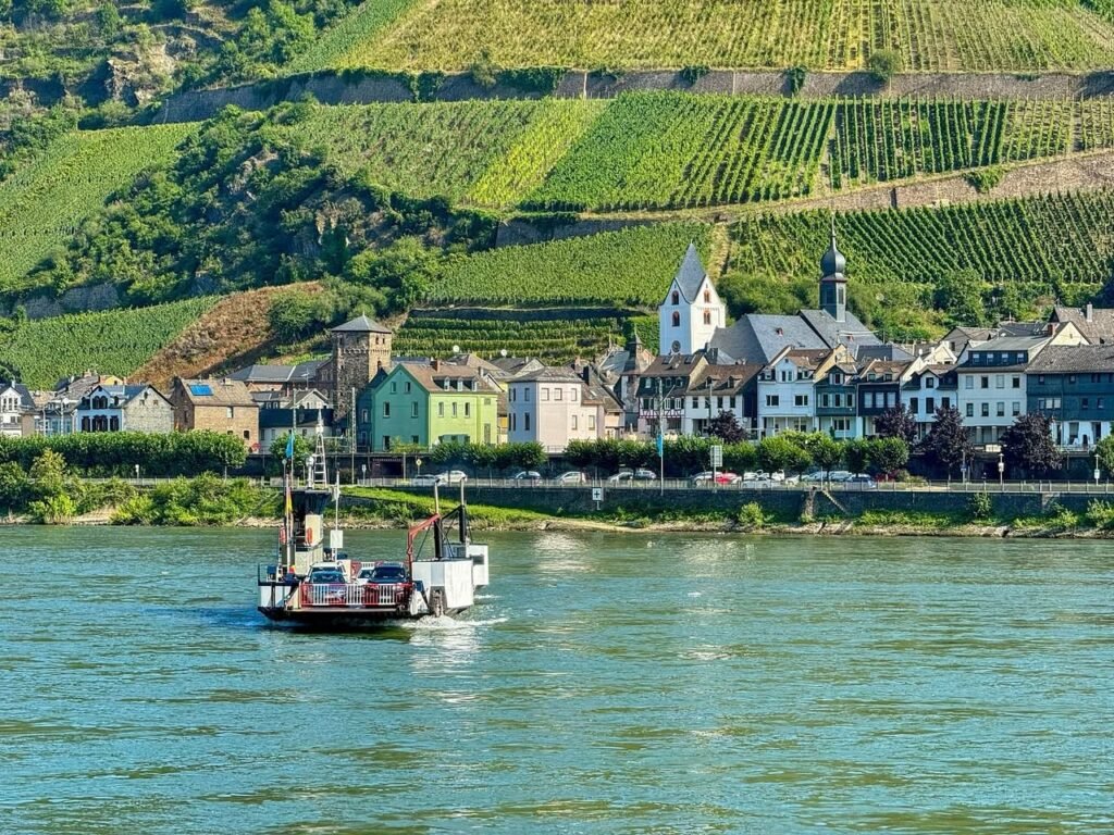 A peaceful river scene featuring a ferry boat with a picturesque village and terraced green vineyards in the background.