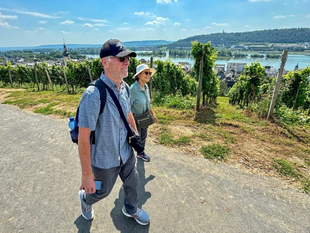 A couple hikes along a paved path overlooking vineyards and a river valley. The landscape features lush greenery and distant hills under a bright blue sky.