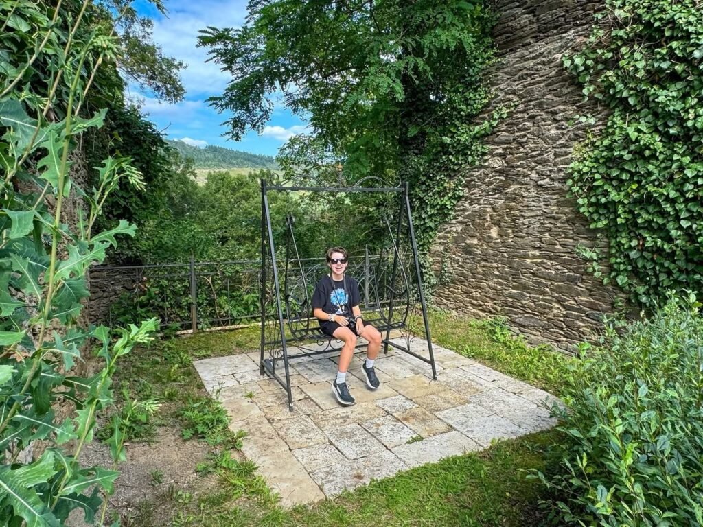 A person enjoys the outdoors on a swing set at a scenic historical site, surrounded by stone walls and greenery.