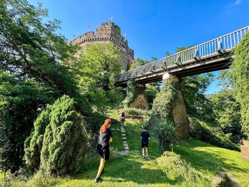 Visitors walk beneath the Fairground Bridge on a sunny day with Edinburgh Castle's iconic tower rising above the green hillside.