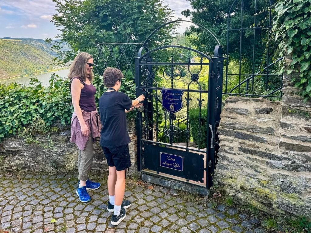 People standing in front of an ornate iron gate. A scenic mountain and river landscape is visible in the background.