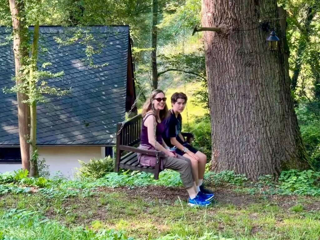 A woman and a man are sitting on a wooden bench next to a lake in a forest. They appear to be enjoying a quiet moment by the water, surrounded by tall trees and greenery.