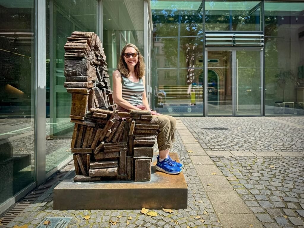 A person sits on a rusted armchair sculpture outside a building. The background features glass panels and pavement.