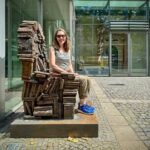 Mainz, Germany A person sits on a rusted armchair sculpture outside a building. The background features glass panels and pavement.