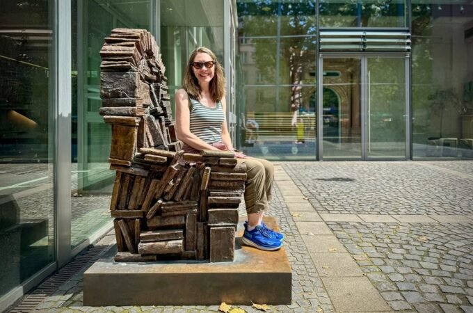 A person sits on a rusted armchair sculpture outside a building. The background features glass panels and pavement.
