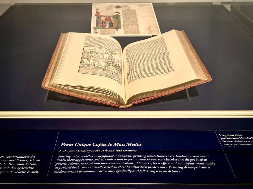 An open ancient book displayed behind glass under museum lighting. The exhibit label reads "From Unique Copies to Mass Media."