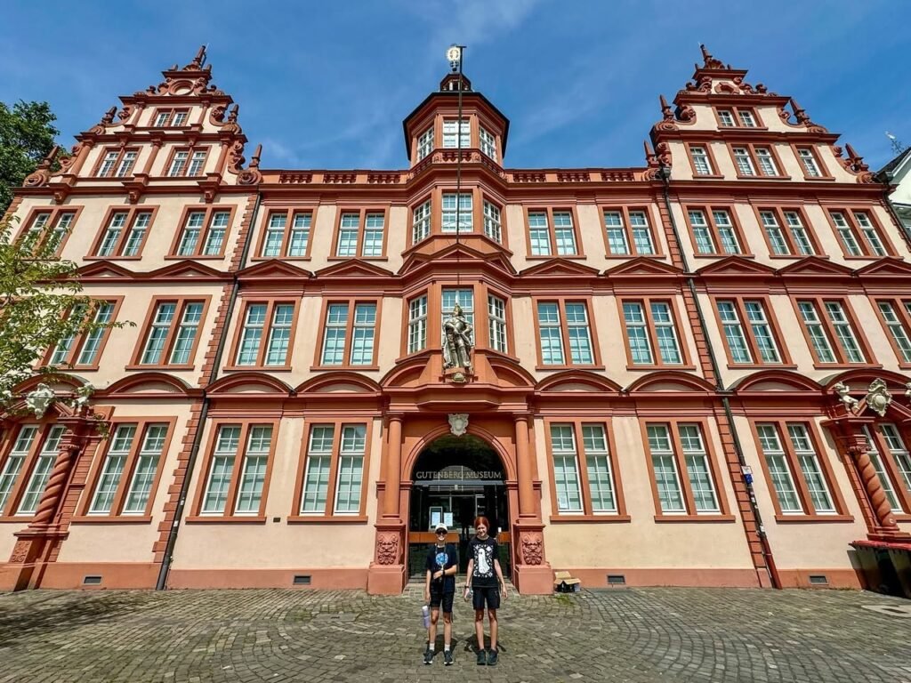 A historic building with ornate red and cream facades. Travelers are walking on a cobblestone courtyard.