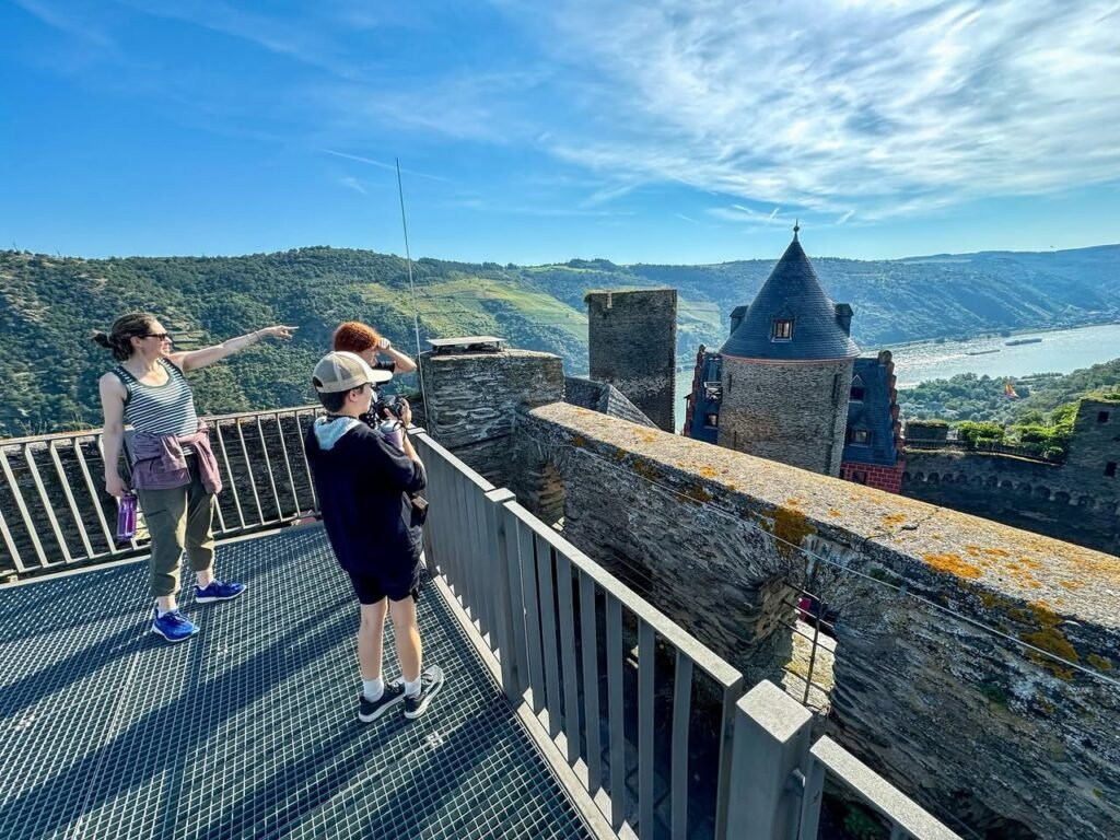 People on a castle rampart overlooking a river valley under a bright blue sky. Stone architecture contrasts with sunshades.