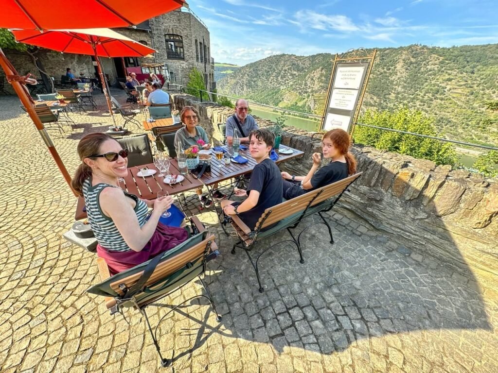 A group of people enjoying a meal at an outdoor dining area with red umbrellas overlooking a scenic valley.