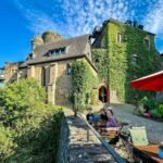 A bright view of an old, ivy-covered stone building with a courtyard, stone wall, greenery, and modern furniture. People relax in the sun under a red patio umbrella on a clear blue day.
