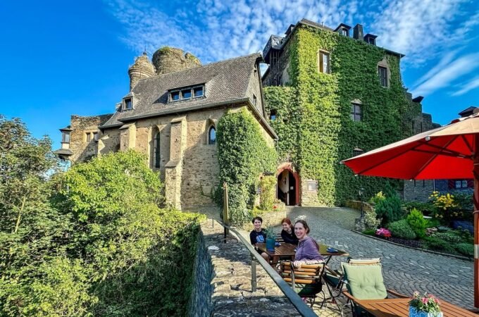 A bright view of an old, ivy-covered stone building with a courtyard, stone wall, greenery, and modern furniture. People relax in the sun under a red patio umbrella on a clear blue day.