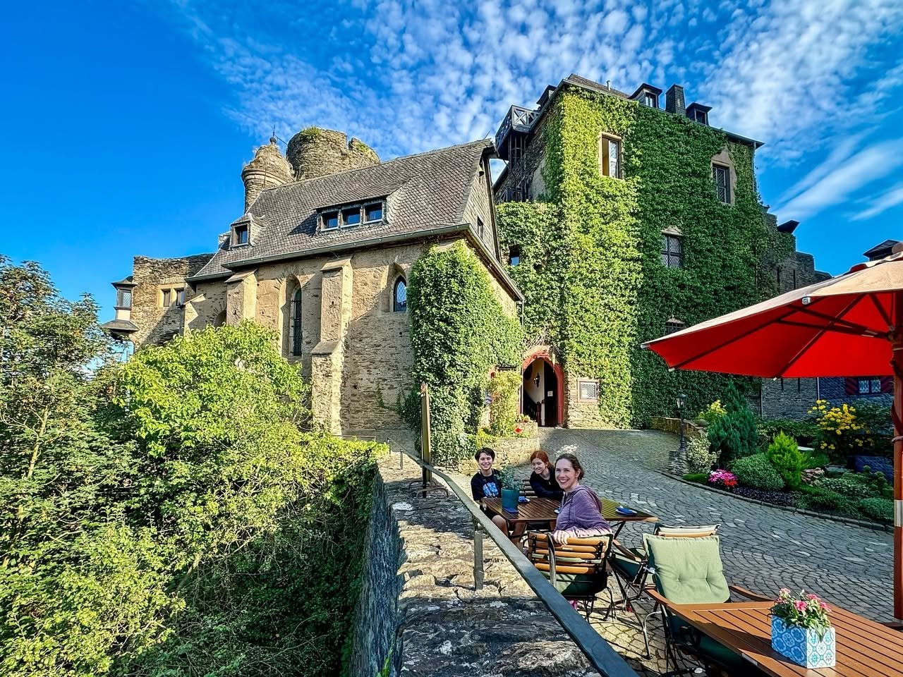 The Castle Museum A bright view of an old, ivy-covered stone building with a courtyard, stone wall, greenery, and modern furniture. People relax in the sun under a red patio umbrella on a clear blue day.