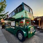 The Castle Museum A classic green double-decker bus parked on a cobblestone street next to a wooden building, with trees and a clear sky in the background.