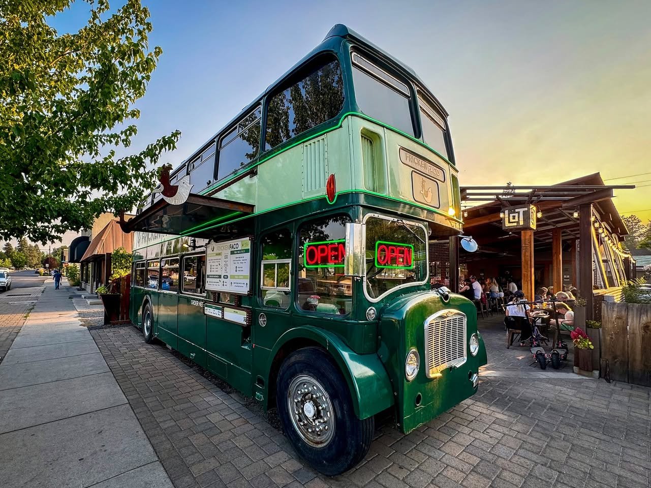 Bend A classic green double-decker bus parked on a cobblestone street next to a wooden building, with trees and a clear sky in the background.