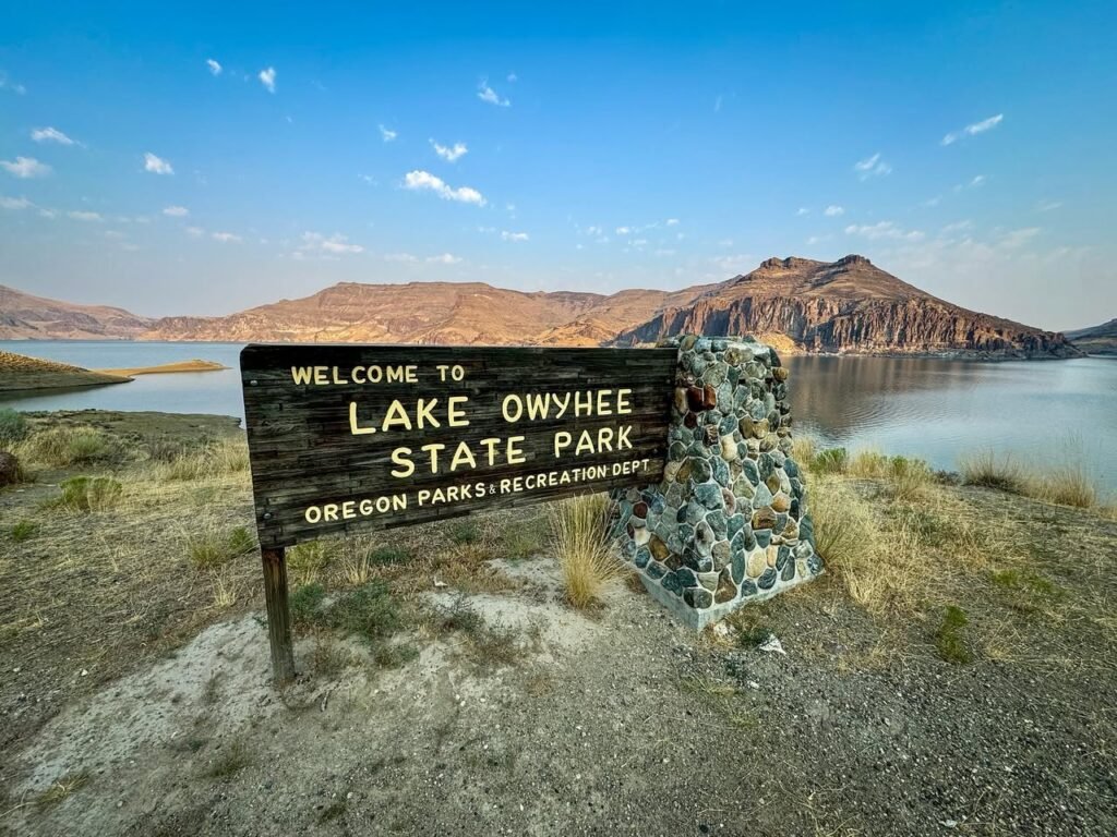 A wooden sign welcoming visitors to Lake Owyhee State Park in Oregon. The scenic view includes the lake, dry desert vegetation, and rugged hills under a clear blue sky.