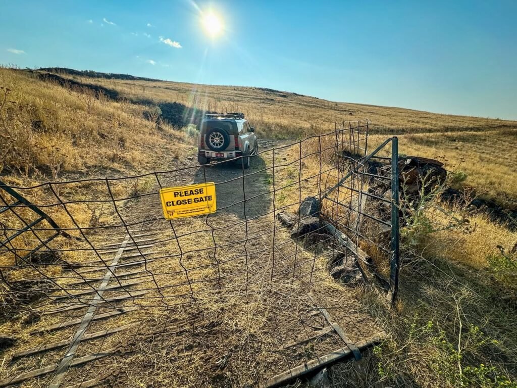 A vehicle navigates a fenced path through dry grasslands under a bright sun with hills in the background.
