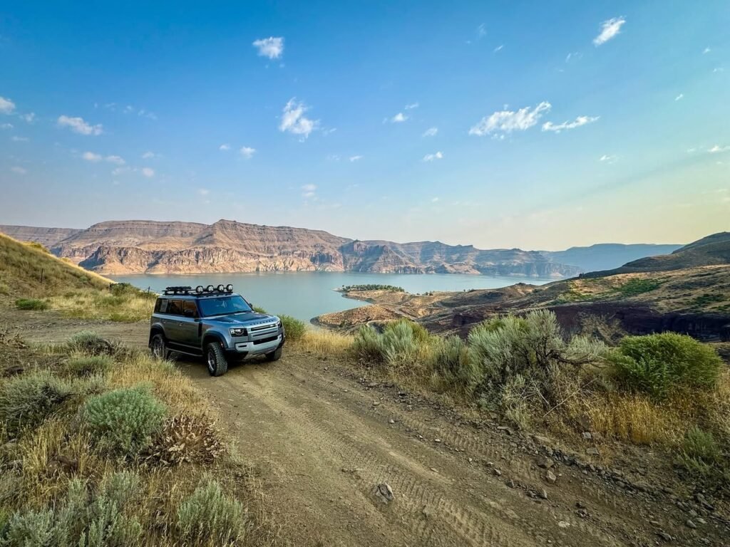 An SUV navigates a rugged dirt trail approaching a scenic overlook of an expansive lake surrounded by rugged hills.
