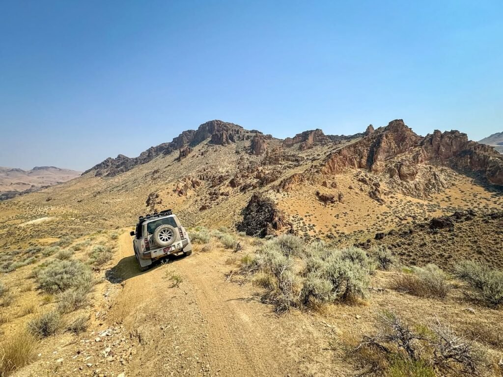 A Jeep travels a dusty trail through a rugged desert landscape with rocky canyons under a clear blue sky.