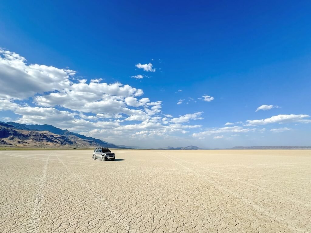 A panoramic view of a car on a dry, cracked desert floor under a bright blue sky with scattered clouds and distant mountains.